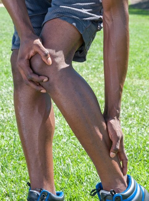 Closeup of lean muscular African American male runner massaging injured leg suffering from Achilles tendonitis as he stands in shorts on green lawn